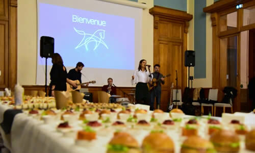 Des petits fours sont disposés sur un buffet au premier plan, on aperçoit une chanteuse accompagnée de musiciens au second plan