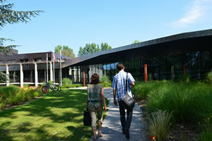 Deux personnes se dirigent sur une allée avec de la pelouse à gauche et des buissons à droite vers un bâtiment bas 
