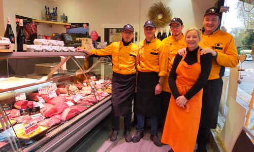 La photo présente 4 hommes et une femme avec des tenues en orange et noires à l'intérieur de la boutique, les bouchers. On y voit également un étal avec différents morceaux de viandes.